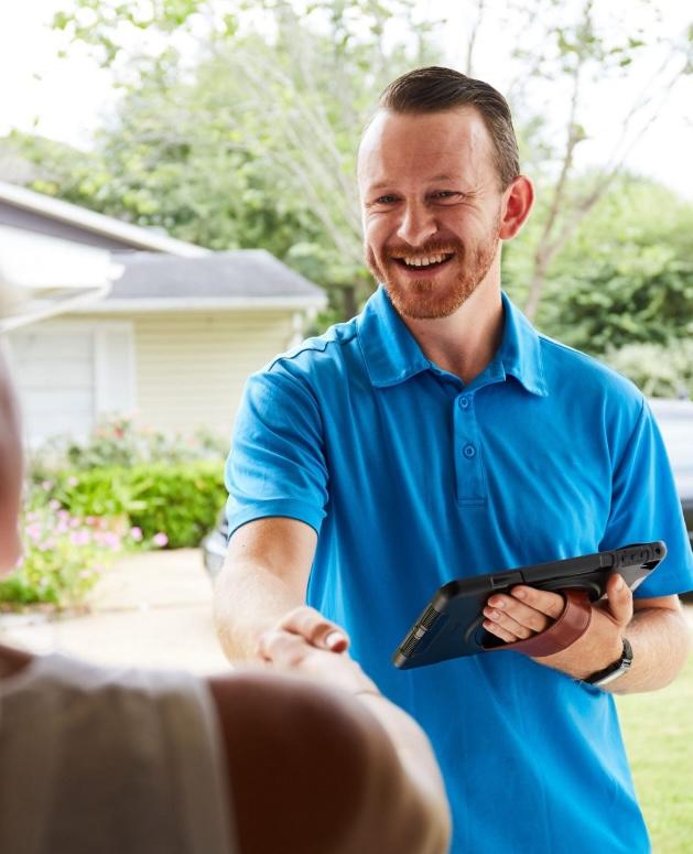 A smiling man in a blue polo shirt, holding a tablet, extends a hand for a handshake with someone off-camera outside a residential setting.