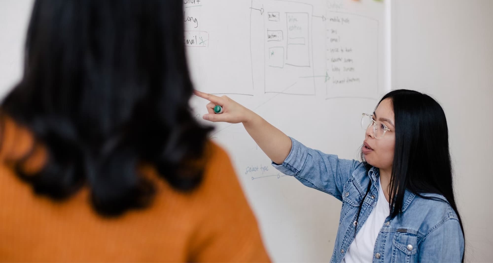 Two women in an office in front of a white board, the woman on the right is pointing to a diagram on the white board