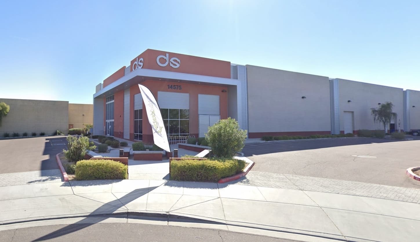 A gray and orange single-story office building, to the right a medium-sized parking lot, and in front a path to the door with some light shrubbery