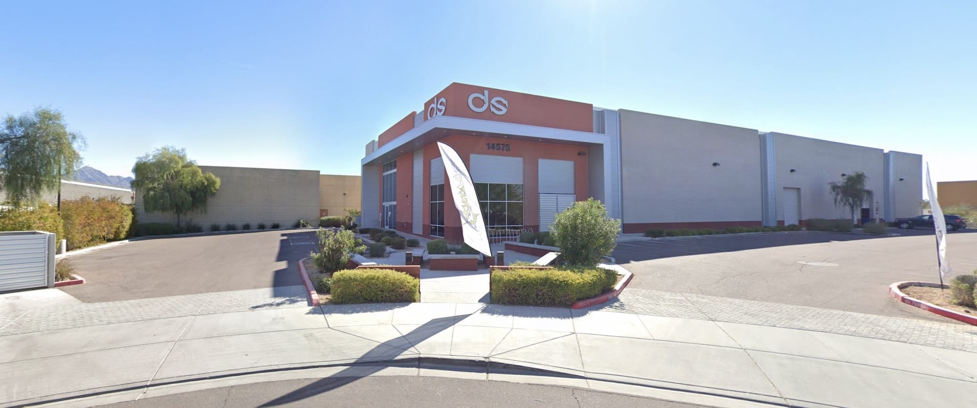 A gray and orange single-story office building, to the right a medium-sized parking lot, and in front a path to the door with some light shrubbery