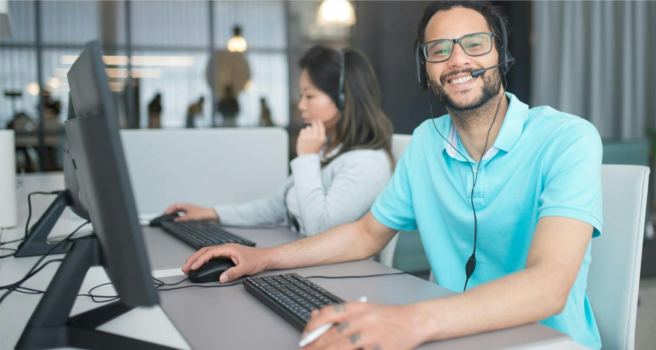 A man in an office wearing glasses and a headset sitting at a computer, smiling at the camera. Behind him a woman, also at a computer, speaking into her headset